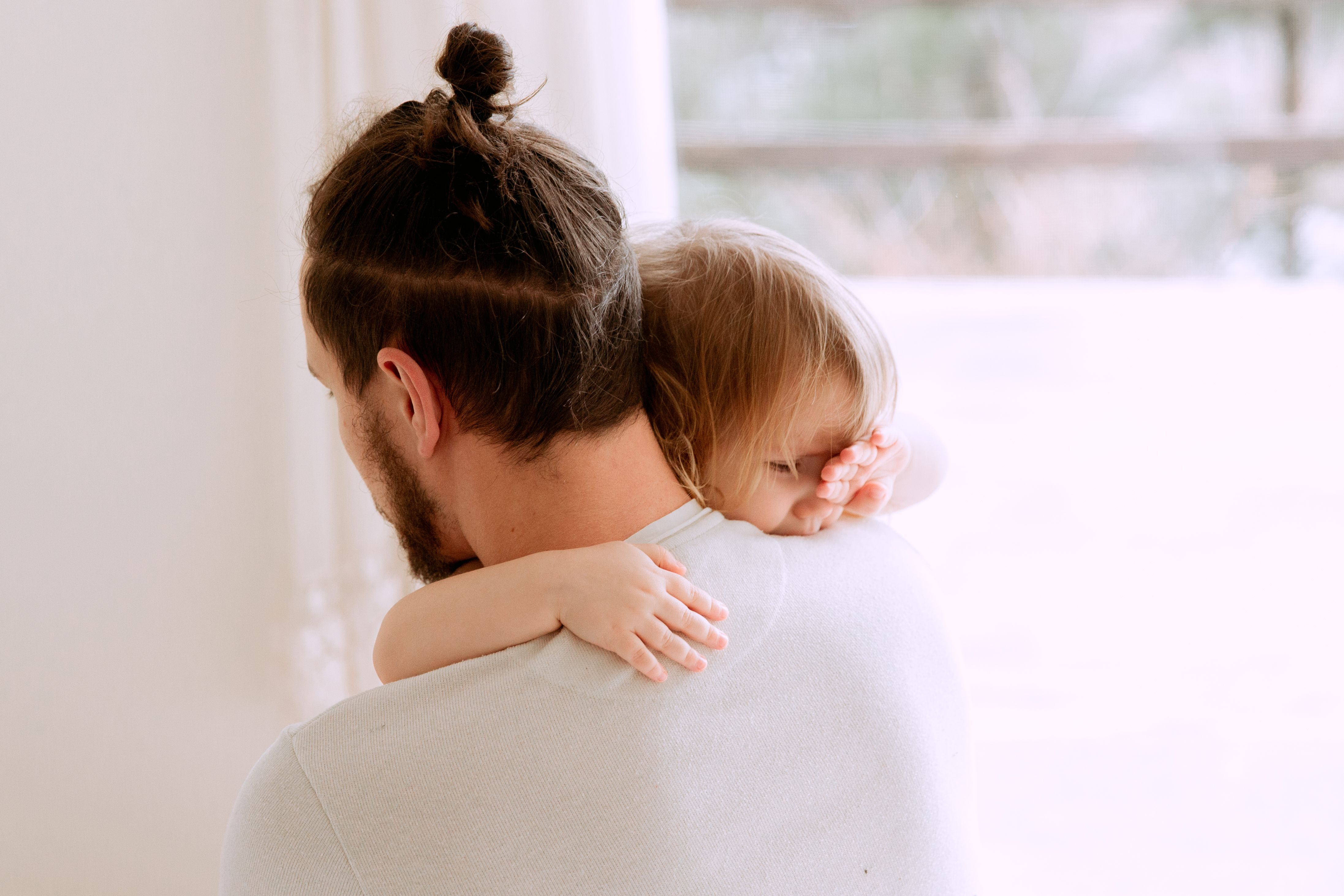 A parent holding a sleeping toddler by a bright window